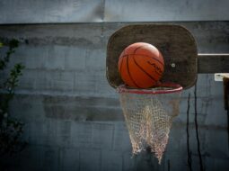 An orange basketball resting on the rim of a rustic outdoor basketball hoop with a wooden backboard and worn net.