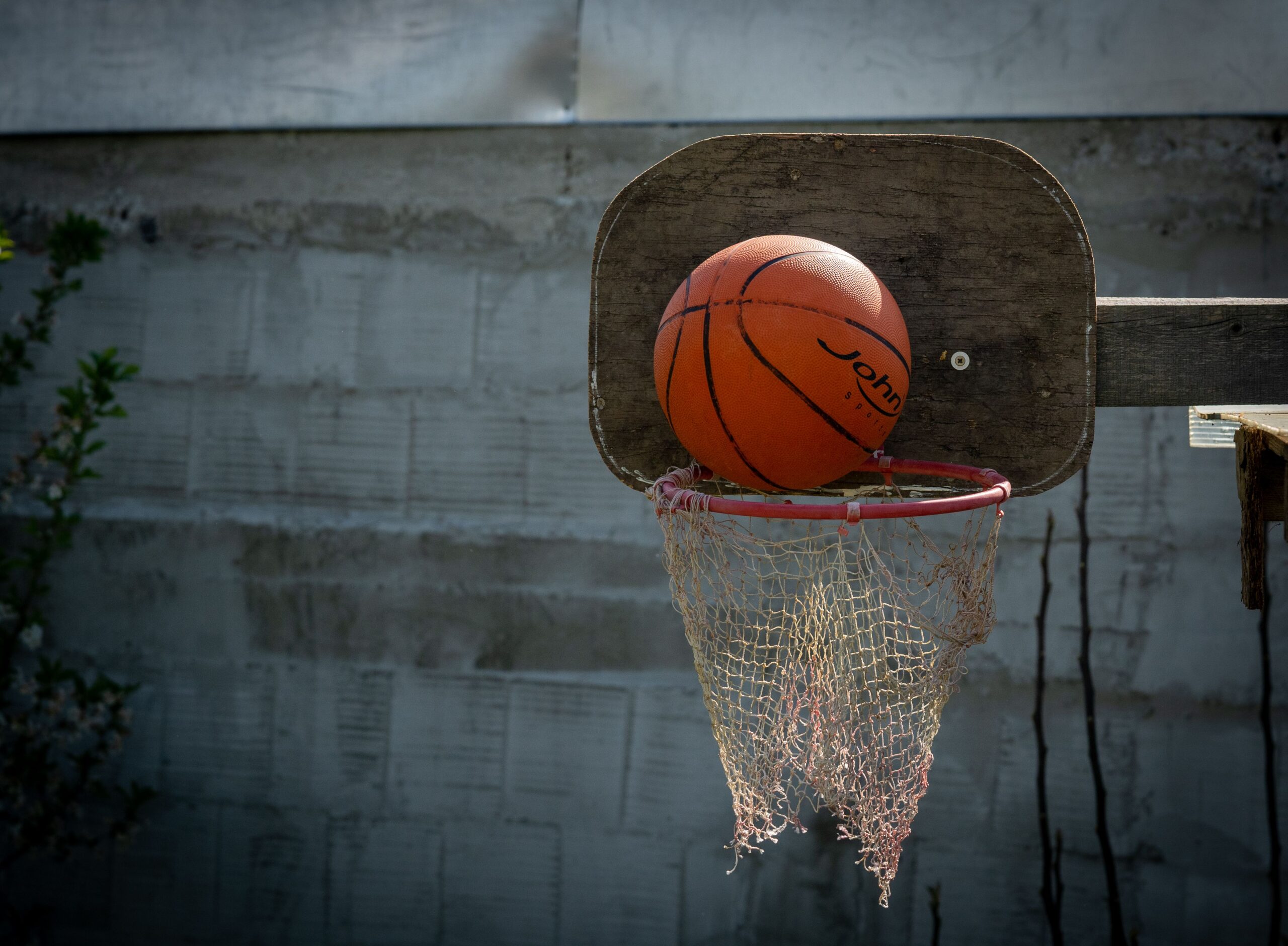 An orange basketball resting on the rim of a rustic outdoor basketball hoop with a wooden backboard and worn net.